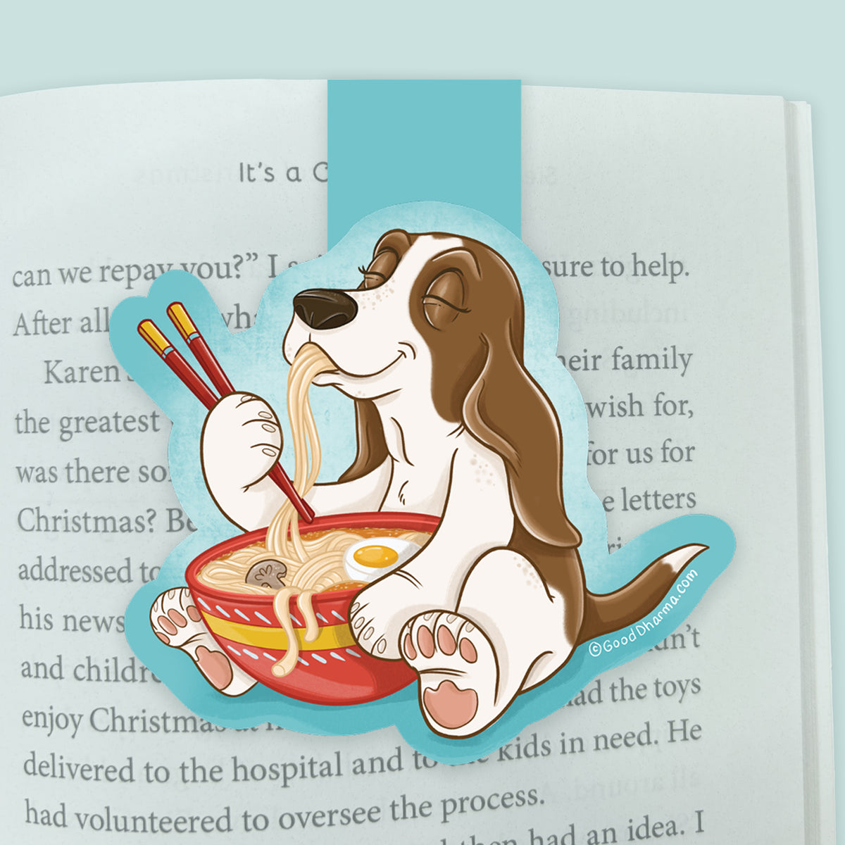 An illustrated magnetic bookmark featuring a cartoon basset hound dog eating ramen noodles with chopsticks on a blue background shown clipped over an open page in a book