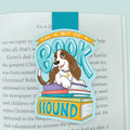 An illustrated magnetic bookmark featuring a cartoon basset hound with a stack of books and hand drawn letters stating 'I'm a bit of a Book Hound' on a light blue background. Shown clipped to a page in an open book