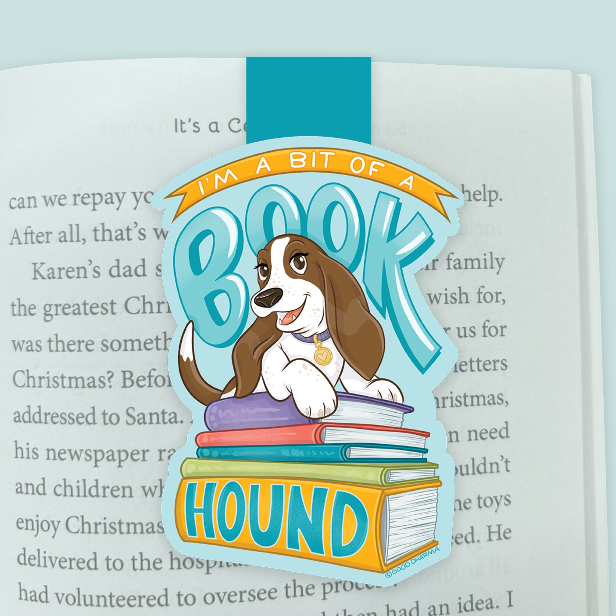 An illustrated magnetic bookmark featuring a cartoon basset hound with a stack of books and hand drawn letters stating 'I'm a bit of a Book Hound' on a light blue background. Shown clipped to a page in an open book