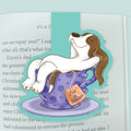 A magnetic bookmark featuring a cartoon dog lounging inside a purple polka-dot teacup with a saucer, clipped to a page in an open book. The bookmark has a turquoise outline and a small orange  tea bag tag reading 'Pup & Saucer'.