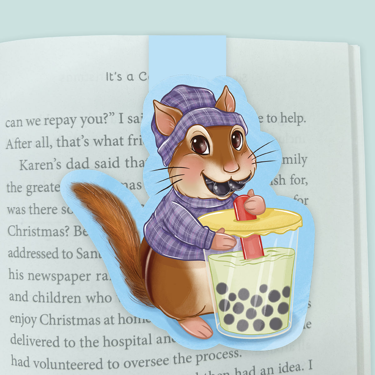 A magnetic bookmark featuring a cartoon chipmunk wearing a purple plaid beanie and sweater, holding a cup of boba tea with a yellow lid, placed on an open book page.