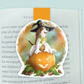 An illustrated magnetic bookmark featuring a cartoon basset hound wearing a witch hat with a spider hanging off o fit while sitting upon a jack-o-lantern pumpkin. Shown clipped to an open page in a book.