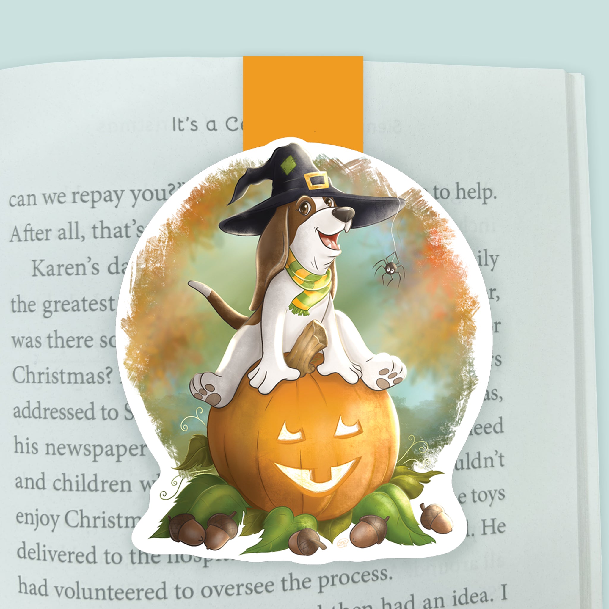 An illustrated magnetic bookmark featuring a cartoon basset hound wearing a witch hat with a spider hanging off o fit while sitting upon a jack-o-lantern pumpkin. Shown clipped to an open page in a book.