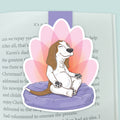 A magnetic bookmarkfeaturing a cartoon basset hound dog sitting cross-legged in yoga lotus pose on a purple cushion, surrounded by a pink lotus flower-like outline, clipped to a page in an open book.