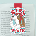 An illustrated magnetic bookmark featuring a basset hound in the Rosie the Riveter pose with text stating 'Girl Power'. Shown clipped to an open page in a book. 