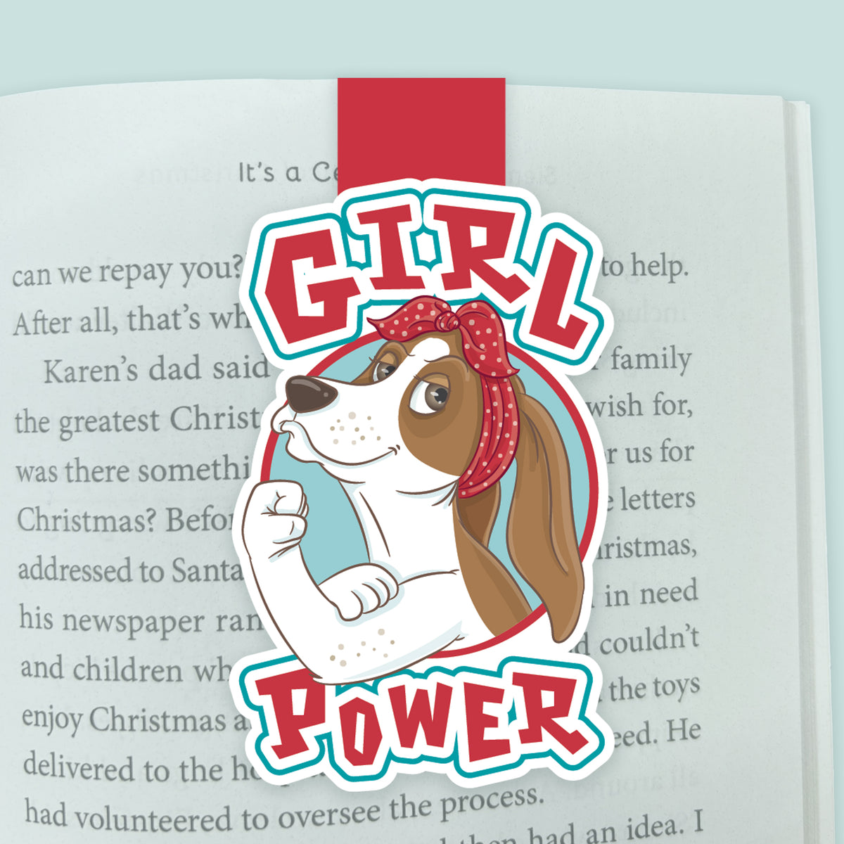 An illustrated magnetic bookmark featuring a basset hound in the Rosie the Riveter pose with text stating 'Girl Power'. Shown clipped to an open page in a book. 