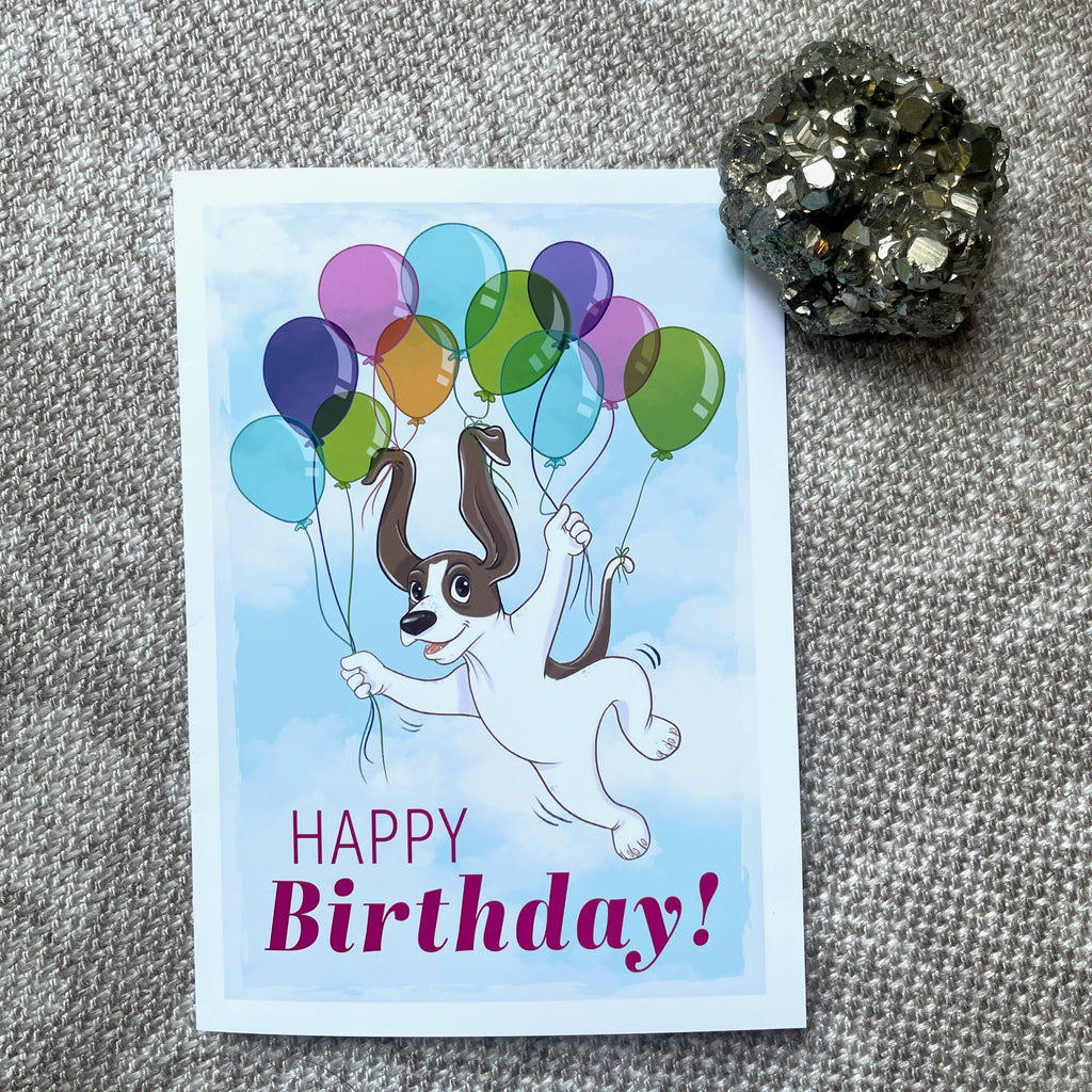 Birthday card with a dog holding balloons on a textured surface.