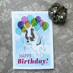 Birthday card with a dog holding balloons on a textured surface.