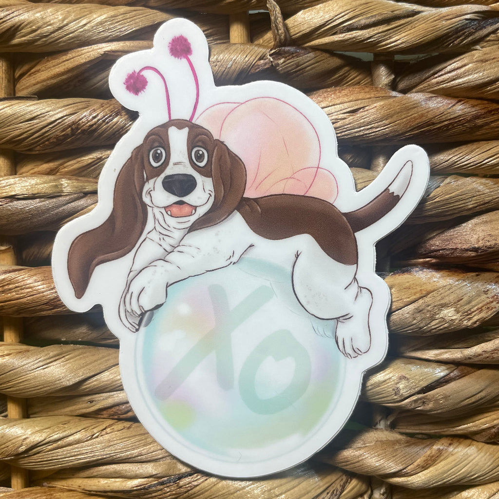 Sticker of a dog with butterfly wings floating on a bubble that says "XO" on a woven surface