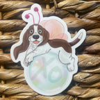 Sticker of a dog with butterfly wings floating on a bubble that says "XO" on a woven surface