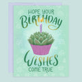 A birthday greeting card with a coordinating purple envelope. The card features a soft green background with white succulent floral motifs, a central illustration of a succulent in a pink cupcake with a lit purple candle, and the text 'HOPE YOUR BIRTHDAY WISHES COME TRUE' in green hand lettering.