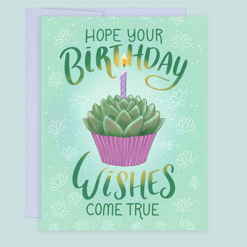 A birthday greeting card with a coordinating purple envelope. The card features a soft green background with white succulent floral motifs, a central illustration of a succulent in a pink cupcake with a lit purple candle, and the text 'HOPE YOUR BIRTHDAY WISHES COME TRUE' in green hand lettering.