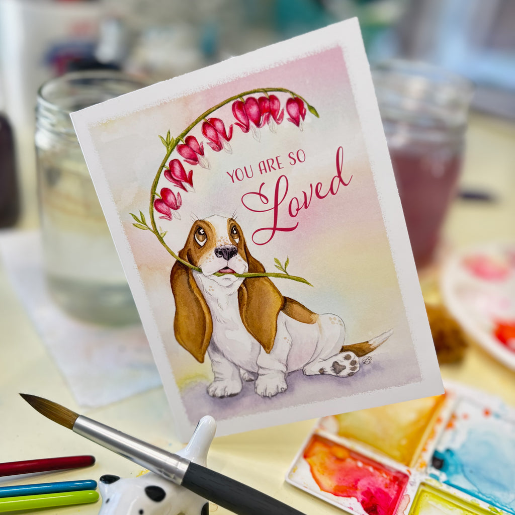 Card with a basset hound holding heart-shaped flowers and 'You are so loved' text, surrounded by art supplies.