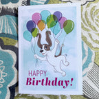Birthday card with a dog holding balloons on a patterned fabric background.