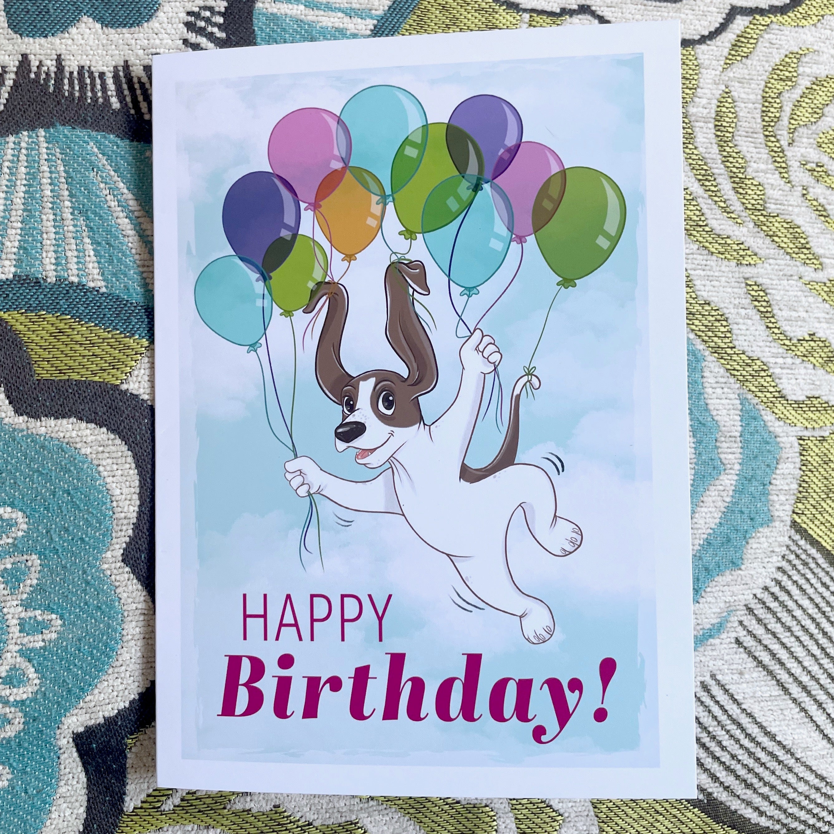 Birthday card with a dog holding balloons on a patterned fabric background.
