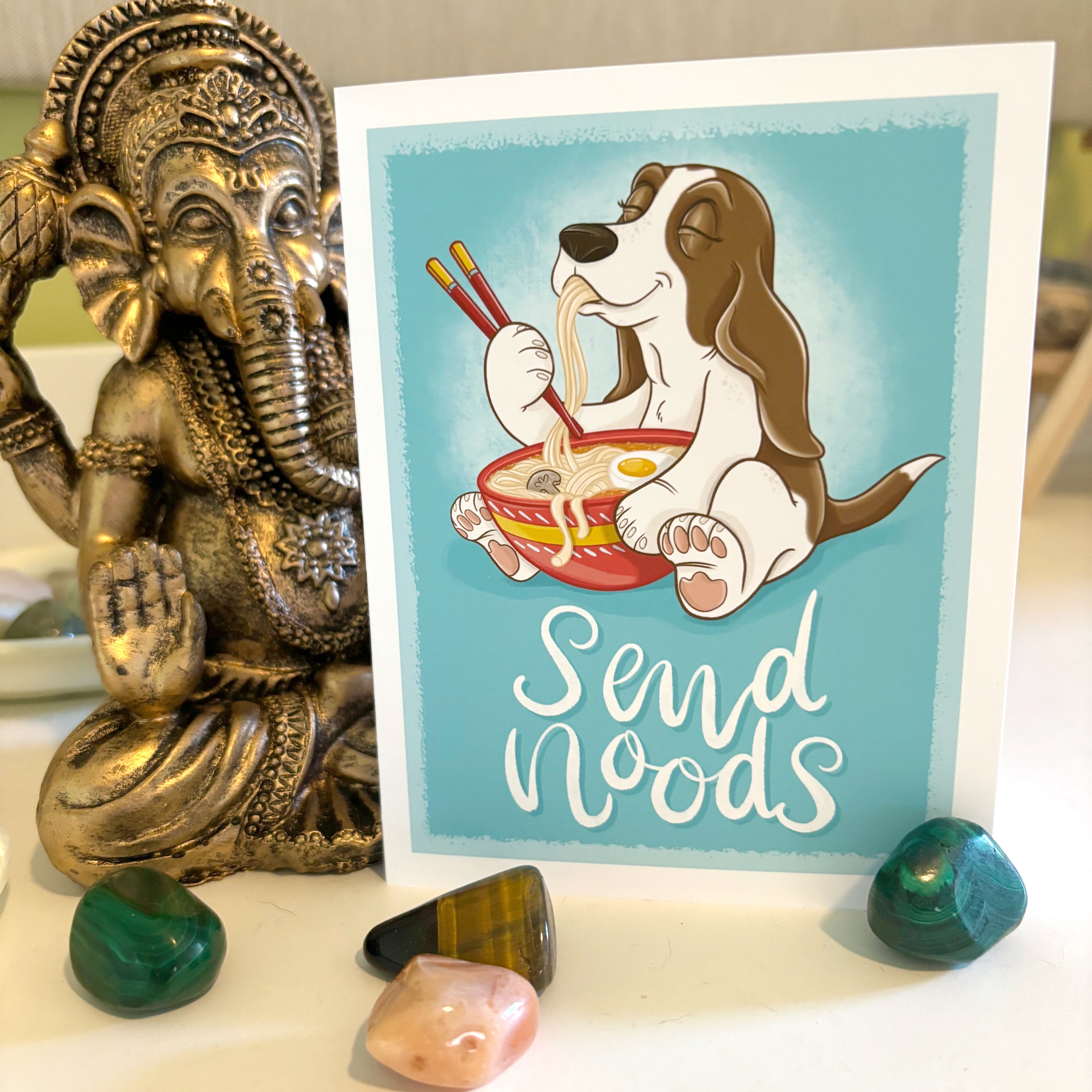 Card with a cartoon dog eating ramen and the text 'Send Noods' next to a decorative statue.