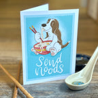 Card with a cartoon dog eating noodles and chopsticks, placed on a wooden surface with a spoon and chopsticks.