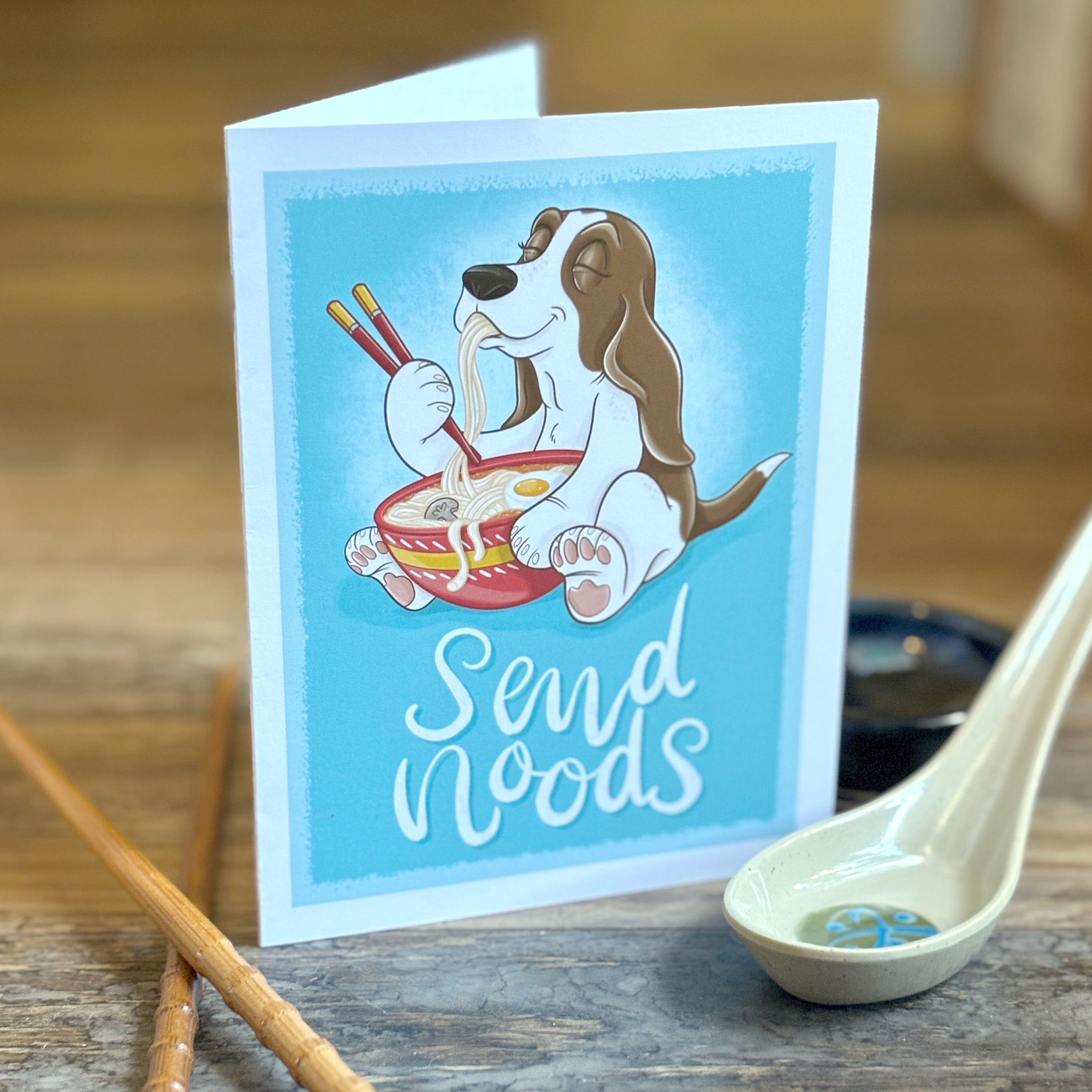 Card with a cartoon dog eating noodles and chopsticks, placed on a wooden surface with a spoon and chopsticks.