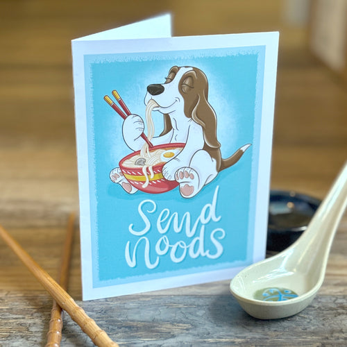 Card with a cartoon dog eating noodles and chopsticks, placed on a wooden surface with a spoon and chopsticks.