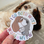 Sticker with dog and cat design and text 'I go both ways woof meow' held by a hand, with a dog in the background.