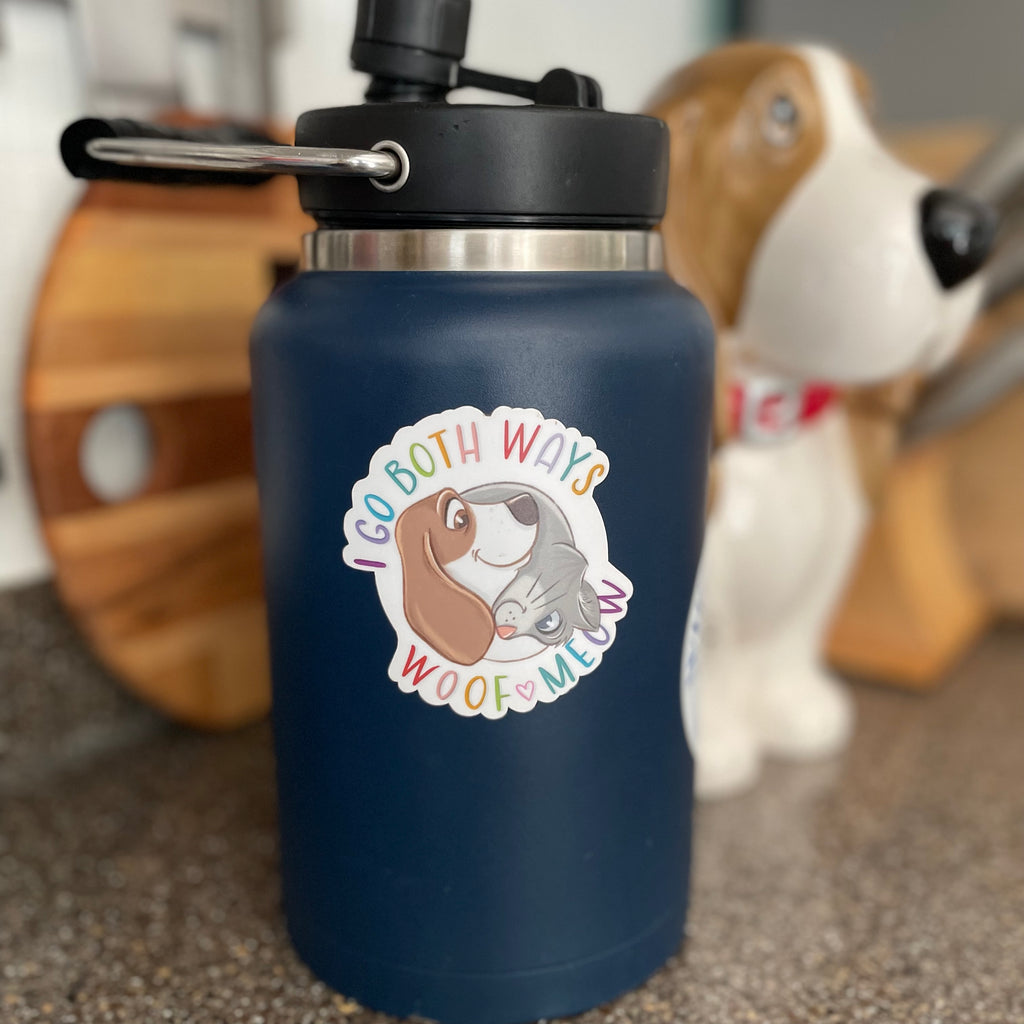 Navy blue water bottle with a colorful sticker featuring a dog and cat, with a blurred background of a wooden barrel and a dog.