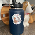 Navy blue water bottle with a colorful sticker featuring a dog and cat, with a blurred background of a wooden barrel and a dog.