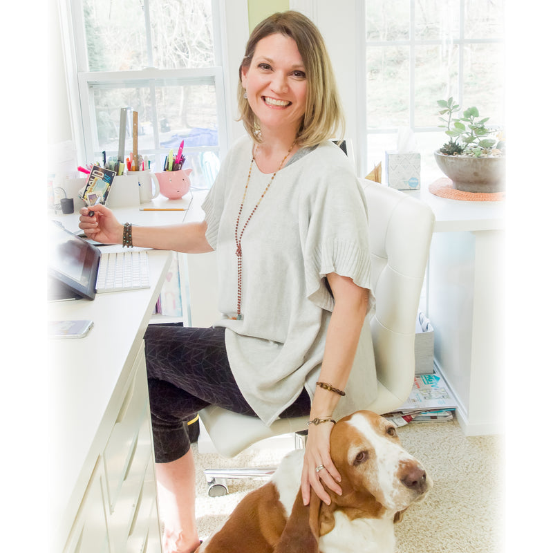 Woman sitting at a desk with a dog on the floor in a bright room.
