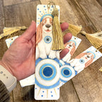 Hand holding a decorative bookmark with a dog illustration and tassel on a wooden surface. ©Good Dharma Design by Zoe Ranucci