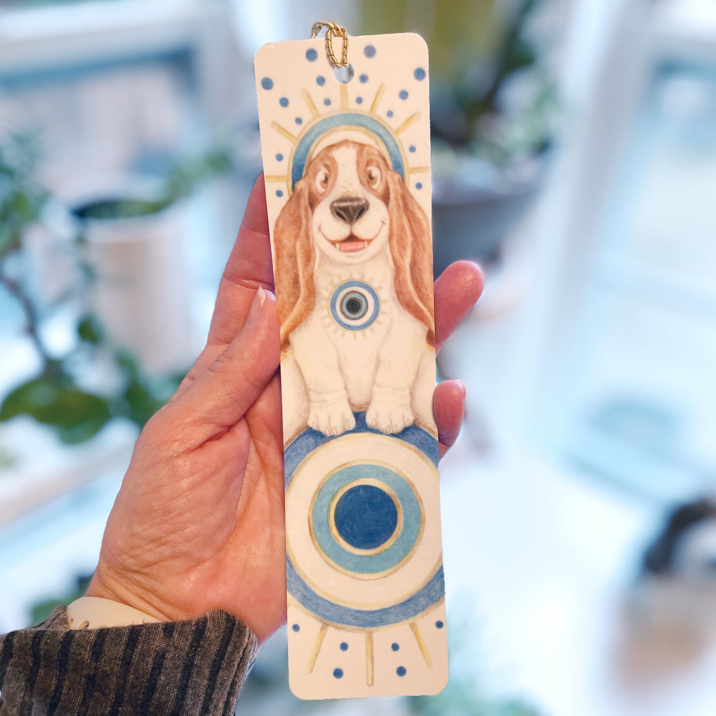 Hand holding a wooden bookmark with a dog illustration against a blurred natural background. ©Good Dharma Design by Zoe Ranucci