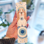 Hand holding a wooden bookmark with a dog illustration against a blurred natural background. ©Good Dharma Design by Zoe Ranucci