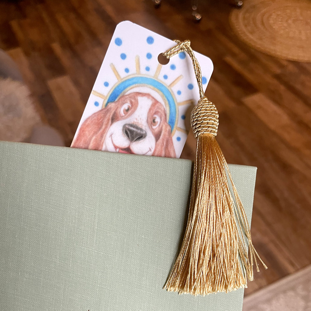 Book with a decorative bookmark featuring a dog illustration and a gold tassel on a wooden surface. ©Good Dharma Design by Zoe Ranucci