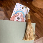 Book with a decorative bookmark featuring a dog illustration and a gold tassel on a wooden surface. ©Good Dharma Design by Zoe Ranucci